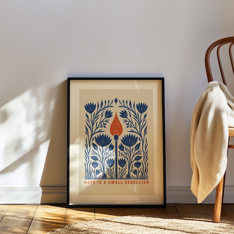 May include: A framed art print featuring a central orange matchstick surrounded by symmetrical blue floral illustrations on a cream background. The text HOPE IS A SMALL REBELLION is printed in orange at the bottom of the rectangular composition.