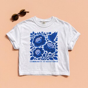 Puede incluir: Una camiseta blanca de manga corta con un estampado gr&aacute;fico azul de tres flores grandes y un p&aacute;jaro. Debajo de la ilustraci&oacute;n, el texto "COMMUNITY IS RESISTANCE" aparece en letras may&uacute;sculas azules. Unas gafas de sol est&aacute;n situadas cerca.