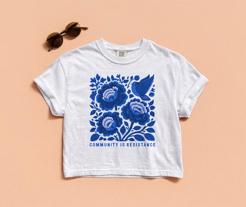 Puede incluir: Una camiseta blanca de manga corta con un estampado gr&aacute;fico azul de tres flores grandes y un p&aacute;jaro. Debajo de la ilustraci&oacute;n, el texto "COMMUNITY IS RESISTANCE" aparece en letras may&uacute;sculas azules. Unas gafas de sol est&aacute;n situadas cerca.