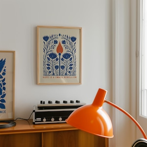 May include: A framed rectangular wall art print featuring a central orange matchstick surrounded by symmetrical blue floral patterns on a light beige background, with the text HOPE IS A SMALL REBELLION printed in orange at the bottom of the print.