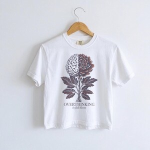 May include: White t-shirt with a graphic of a brain and flower design. The design is split down the middle, with a brain on the left and a flower on the right. The text "OVERTHINKING in full bloom" is below the design.