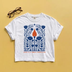 White Comfort Colors boxy crop tee laid flat featuring an original folk art illustration of a lit match rising through dense botanical wildflowers in cobalt blue with red-orange flame, Hope Is A Small Rebellion — activist feminist boxy crop tee.