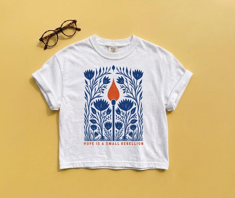 White Comfort Colors boxy crop tee laid flat featuring an original folk art illustration of a lit match rising through dense botanical wildflowers in cobalt blue with red-orange flame, Hope Is A Small Rebellion — activist feminist boxy crop tee.