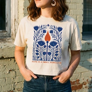 May include: A cream-colored short-sleeve t-shirt featuring a graphic print of blue flowers surrounding a central orange matchstick. Below the illustration, the text reads HOPE IS A SMALL REBELLION in orange capital letters on the front of the shirt.