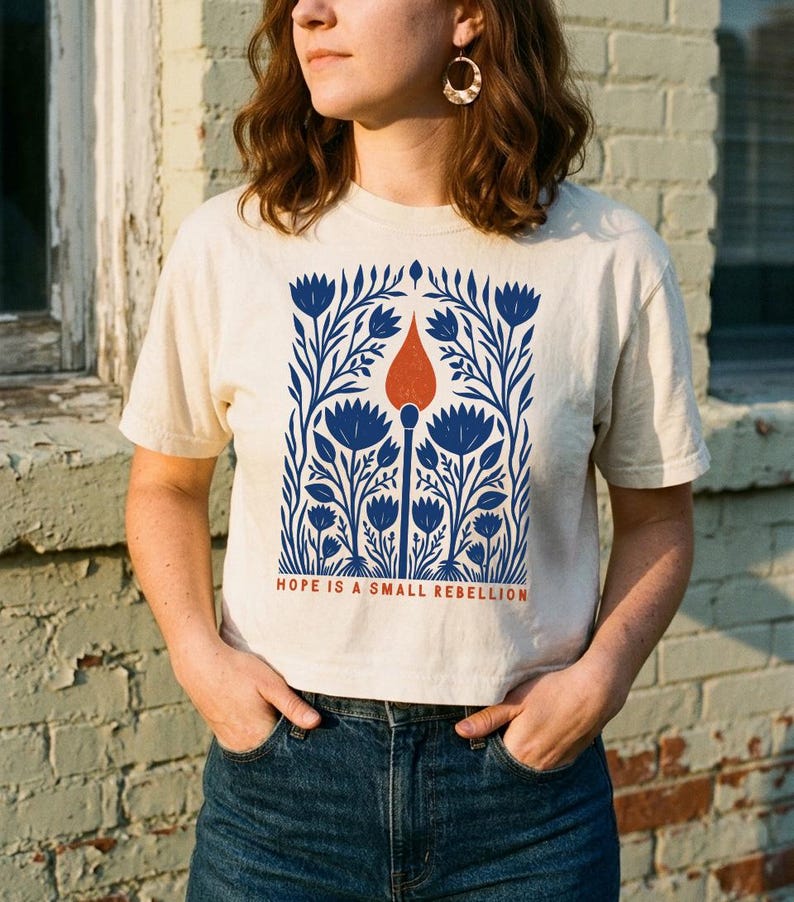 May include: A cream-colored short-sleeve t-shirt featuring a graphic print of blue flowers surrounding a central orange matchstick. Below the illustration, the text reads HOPE IS A SMALL REBELLION in orange capital letters on the front of the shirt.