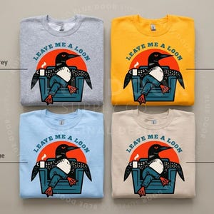 May include: Four crewneck sweatshirts in sport grey, gold, light blue, and sand colors. Each features a graphic of a loon sitting in a chair with a coffee mug, wearing blue jeans, and the text LEAVE ME A LOON printed in a circular arc above.