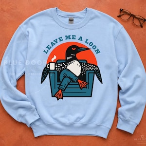 Lake life sweatshirt in light blue styled with vintage glasses on rust background, funny loon graphic crewneck for birders and cozy lake gift shopping