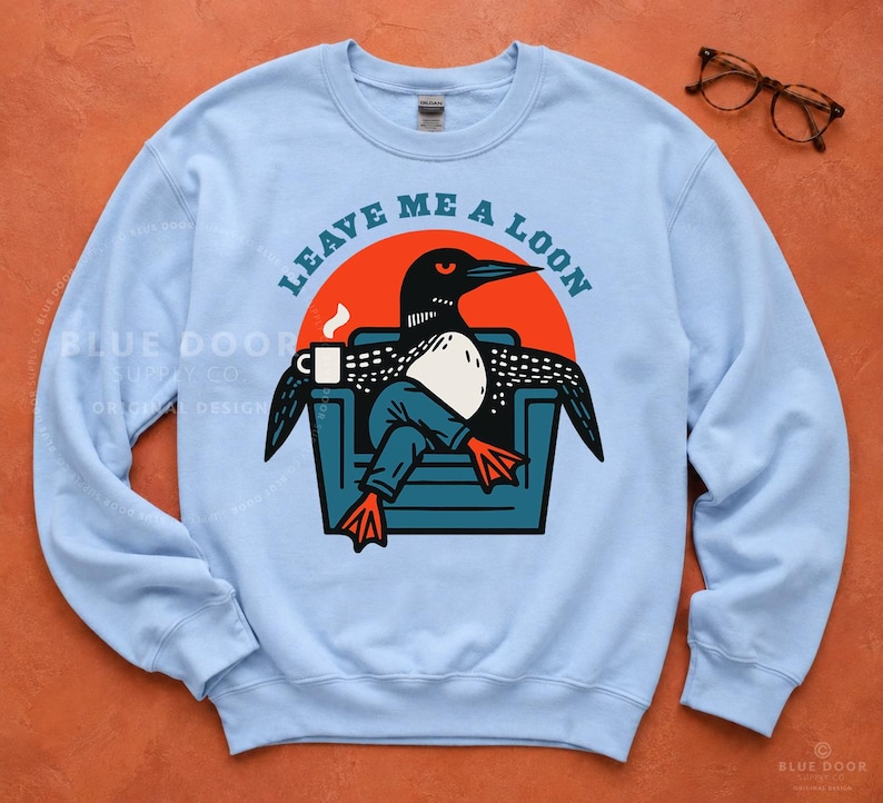Lake life sweatshirt in light blue styled with vintage glasses on rust background, funny loon graphic crewneck for birders and cozy lake gift shopping