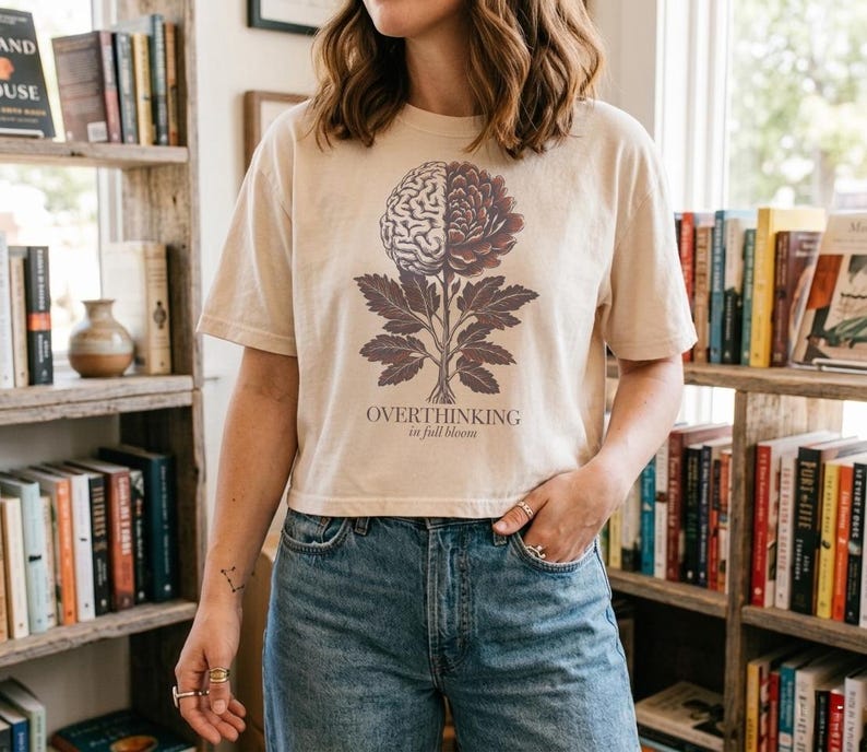 May include: Beige t-shirt with a graphic of a brain-flower and the text "OVERTHINKING in full bloom". The design is in shades of brown and purple. The shirt is paired with blue jeans.
