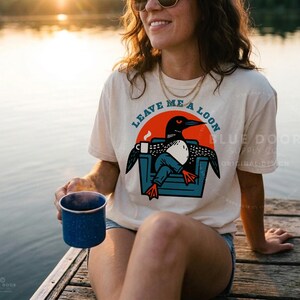 May include: A cream-colored short-sleeve t-shirt featuring a graphic of a loon sitting in a blue chair while holding a white coffee mug. The text "LEAVE ME A LOON" is printed in an arc above the graphic, which is set against a solid orange circle.