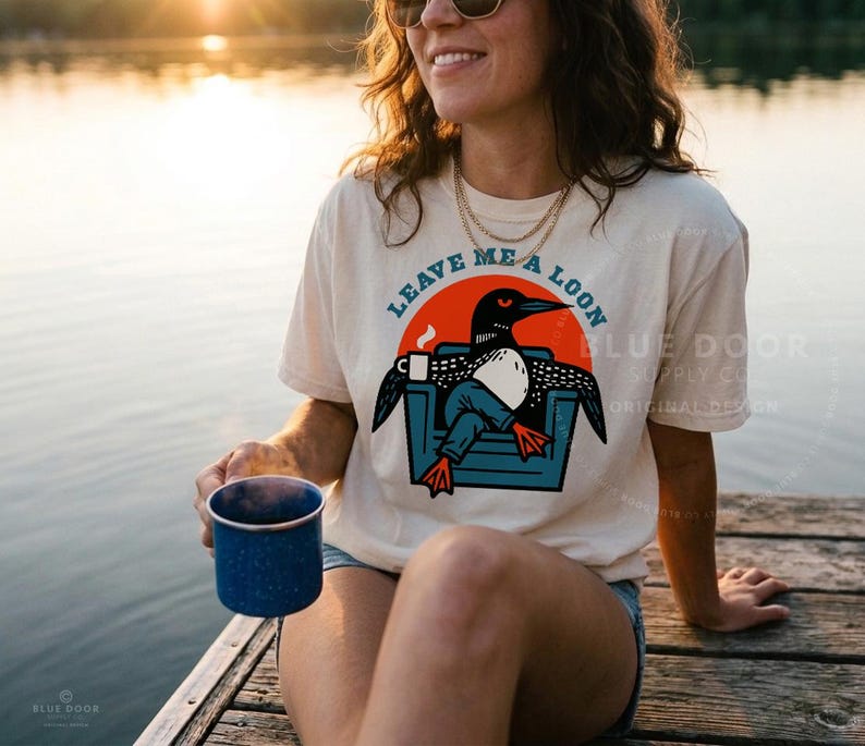 May include: A cream-colored short-sleeve t-shirt featuring a graphic of a loon sitting in a blue chair while holding a white coffee mug. The text "LEAVE ME A LOON" is printed in an arc above the graphic, which is set against a solid orange circle.