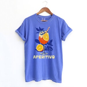 May include: A Flo-blue comfort colors t-shirt with a graphic of an Aperitivo cocktail. The cocktail is in a stemmed glass with an orange slice and a straw. The word "APERITIVO" is printed in yellow below the glass.