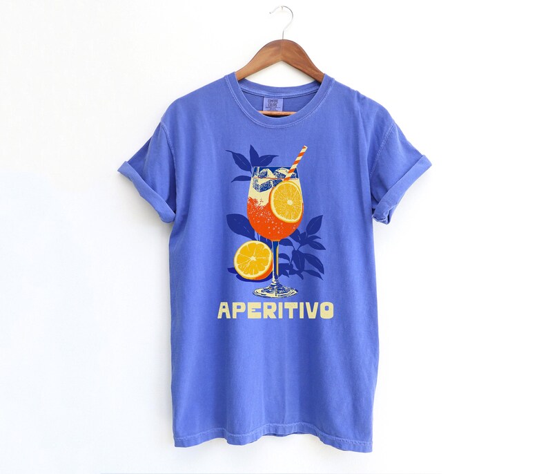May include: A Flo-blue comfort colors t-shirt with a graphic of an Aperitivo cocktail. The cocktail is in a stemmed glass with an orange slice and a straw. The word "APERITIVO" is printed in yellow below the glass.