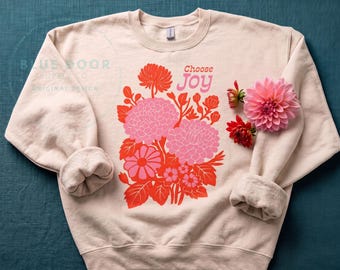 May include: A light beige crewneck sweatshirt featuring a graphic print of pink and orange flowers with the text "Choose Joy" in pink and red. The design includes stylized floral illustrations and is accented by fresh pink and red dahlia flowers.