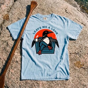 Loon gift shirt in chambray blue laid on a lakeside rock beside a wooden canoe paddle, camp-core funny loon graphic tee for outdoor and lake house enthusiasts, retro bird lover apparel