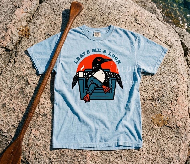 Loon gift shirt in chambray blue laid on a lakeside rock beside a wooden canoe paddle, camp-core funny loon graphic tee for outdoor and lake house enthusiasts, retro bird lover apparel