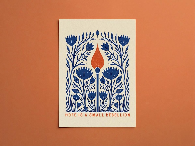 May include: A rectangular print on cream paper featuring a central blue matchstick with an orange flame, surrounded by symmetrical blue floral and leaf patterns. The text HOPE IS A SMALL REBELLION is printed in orange at the bottom of the design.
