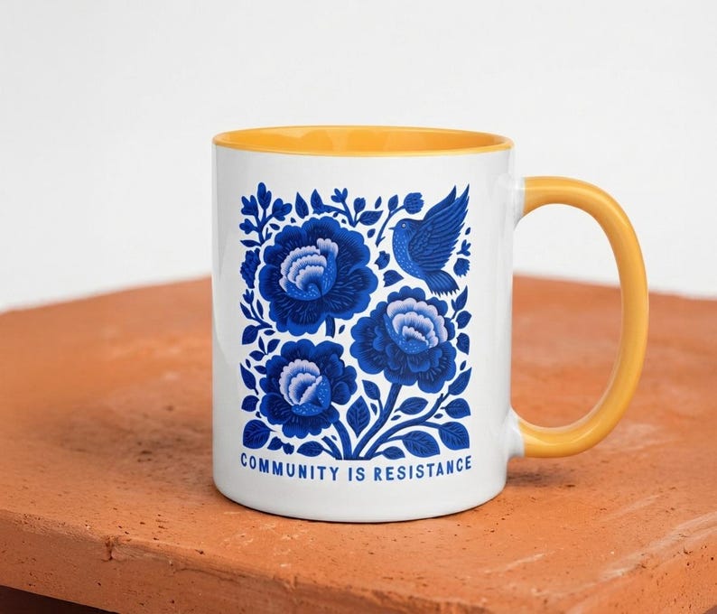 May include: White ceramic mug with a yellow handle and interior. The mug features a blue floral design with a bird and the text "COMMUNITY IS RESISTANCE". The mug is sitting on a terracotta surface.