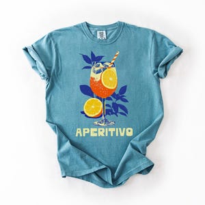 May include: A turquoise t-shirt features a graphic of an Aperitivo cocktail with orange slices, ice, and a straw. The design includes blue leaves and the word "APERITIVO" in yellow.