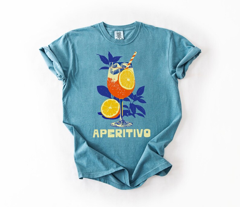 May include: A turquoise t-shirt features a graphic of an Aperitivo cocktail with orange slices, ice, and a straw. The design includes blue leaves and the word "APERITIVO" in yellow.