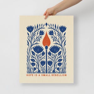 Hope Is a Small Rebellion PosterFolk art print of a lit match rising through cobalt blue wildflowers with a red-orange flame on cream, text reading Hope Is A Small Rebellion. A resistance poster and activism art print with a hand-carved block print look.