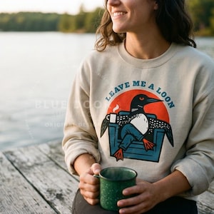 oman standing at a lake dock wearing a sand loon sweatshirt and holding a coffee mug, funny Leave Me A Loon crewneck for cabin and lake life gifting