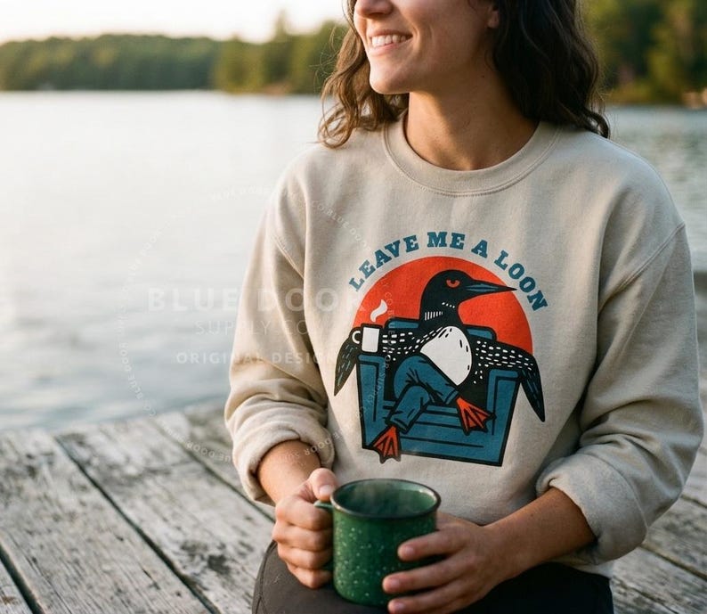 oman standing at a lake dock wearing a sand loon sweatshirt and holding a coffee mug, funny Leave Me A Loon crewneck for cabin and lake life gifting