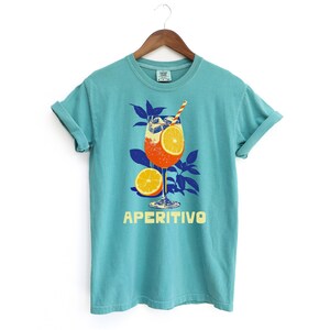 May include: A turquoise t-shirt with a graphic of an Aperitivo cocktail, featuring orange slices and a straw. The word "APERITIVO" is printed in yellow below the drink illustration. The shirt is hanging on a wooden hanger.