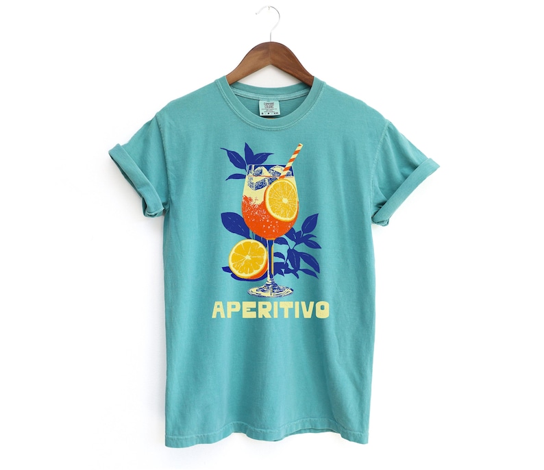 May include: A turquoise t-shirt with a graphic of an Aperitivo cocktail, featuring orange slices and a straw. The word "APERITIVO" is printed in yellow below the drink illustration. The shirt is hanging on a wooden hanger.