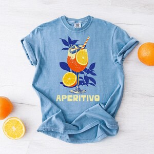 Light blue t-shirt featuring a graphic of an Italian cocktail with orange slices and a straw. The word "APERITIVO" is printed in yellow. Oranges and a slice of orange are placed around the shirt.