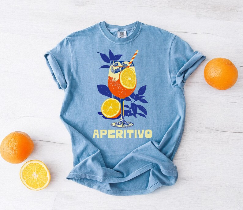Light blue t-shirt featuring a graphic of an Italian cocktail with orange slices and a straw. The word "APERITIVO" is printed in yellow. Oranges and a slice of orange are placed around the shirt.