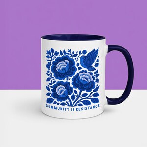 May include: A white ceramic mug with a dark blue interior and handle. The exterior features a blue floral pattern with a bird and the text "COMMUNITY IS RESISTANCE" printed in blue at the bottom. The design is centered on the front of the mug.