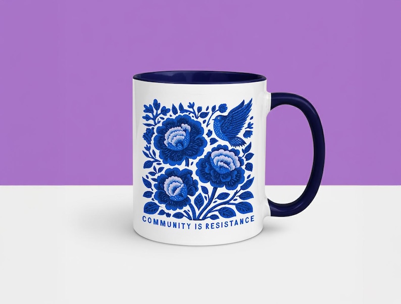 May include: A white ceramic mug with a dark blue interior and handle. The exterior features a blue floral pattern with a bird and the text "COMMUNITY IS RESISTANCE" printed in blue at the bottom. The design is centered on the front of the mug.
