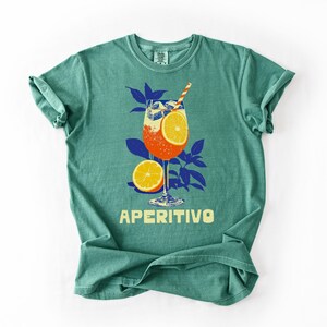 A light green comfort colors t-shirt featuring a graphic of an Aperitivo cocktail with an orange slice, a straw, and the word "APERITIVO" in yellow. The design includes blue leaves and a half orange.