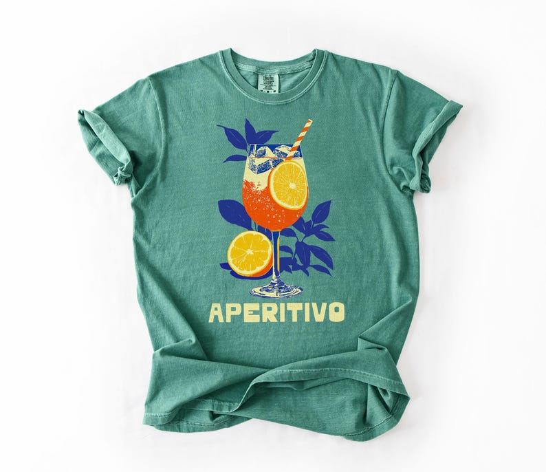 A light green comfort colors t-shirt featuring a graphic of an Aperitivo cocktail with an orange slice, a straw, and the word "APERITIVO" in yellow. The design includes blue leaves and a half orange.