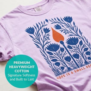 May include: A light purple t-shirt featuring a graphic print of blue flowers surrounding a single orange matchstick. The text "HOPE IS A SMALL RE" is printed in orange at the bottom right. A teal circular overlay contains the text "PREMIUM HEAVYWEIGHT COTTON Signature Softness and Built to Last".