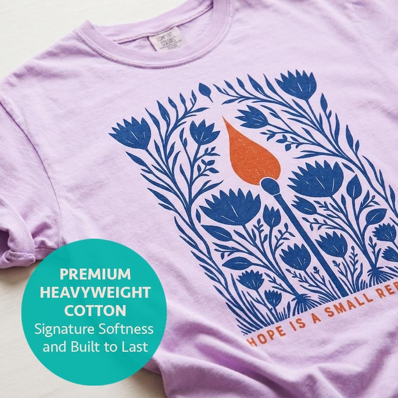 May include: A light purple t-shirt featuring a graphic print of blue flowers surrounding a single orange matchstick. The text "HOPE IS A SMALL RE" is printed in orange at the bottom right. A teal circular overlay contains the text "PREMIUM HEAVYWEIGHT COTTON Signature Softness and Built to Last".