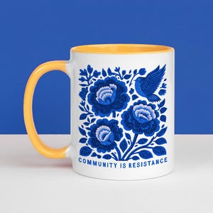May include: A white ceramic mug with a yellow handle and interior. The exterior features a blue floral pattern with a bird and the text "COMMUNITY IS RESISTANCE" printed in blue at the bottom. The design is centered on the front of the mug.