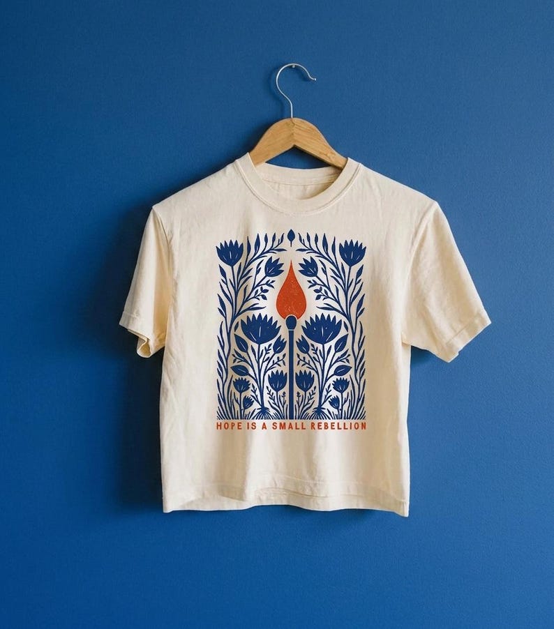 May include: A cream-colored short-sleeve t-shirt featuring a central graphic of a lit match surrounded by blue floral patterns. Below the illustration, the text reads "HOPE IS A SMALL REBELLION" in orange capital letters. The shirt hangs on a wooden hanger.