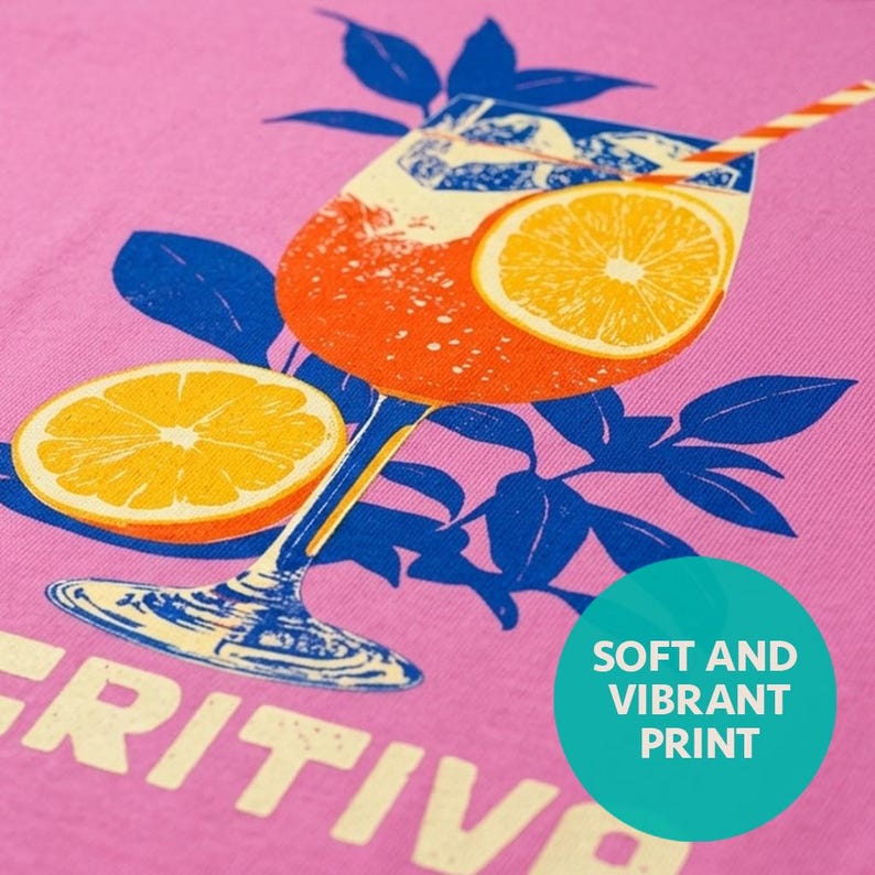 Close-up of Aperitivo graphic tee print on pink — vintage Italian linocut illustration of a spritz cocktail with orange slice, bold blue and orange on a soft women's fitted tee