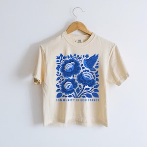Puede incluir: Una camiseta de manga corta de color crema con un estampado gr&aacute;fico azul de tres flores grandes y un p&aacute;jaro. El texto "COMMUNITY IS RESISTANCE" est&aacute; impreso en letras may&uacute;sculas azules debajo del dise&ntilde;o floral. La camiseta cuelga de una percha.