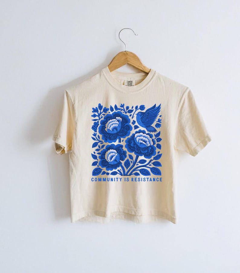 Puede incluir: Una camiseta de manga corta de color crema con un estampado gr&aacute;fico azul de tres flores grandes y un p&aacute;jaro. El texto "COMMUNITY IS RESISTANCE" est&aacute; impreso en letras may&uacute;sculas azules debajo del dise&ntilde;o floral. La camiseta cuelga de una percha.