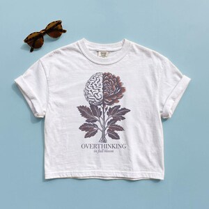 May include: White t-shirt with a graphic of a brain and flower, with the text "OVERTHINKING in full bloom". The shirt is paired with brown tortoise shell sunglasses. The shirt is on a light blue background.