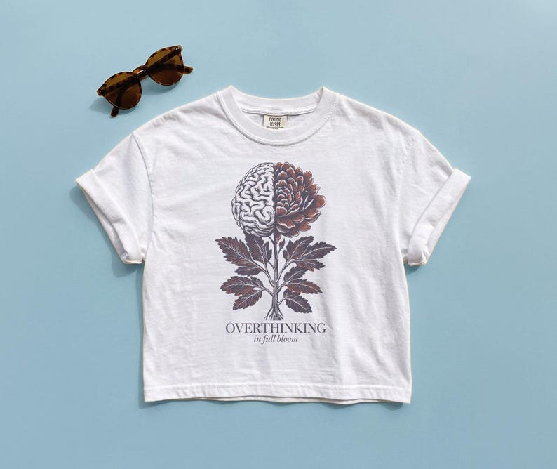 May include: White t-shirt with a graphic of a brain and flower, with the text "OVERTHINKING in full bloom". The shirt is paired with brown tortoise shell sunglasses. The shirt is on a light blue background.