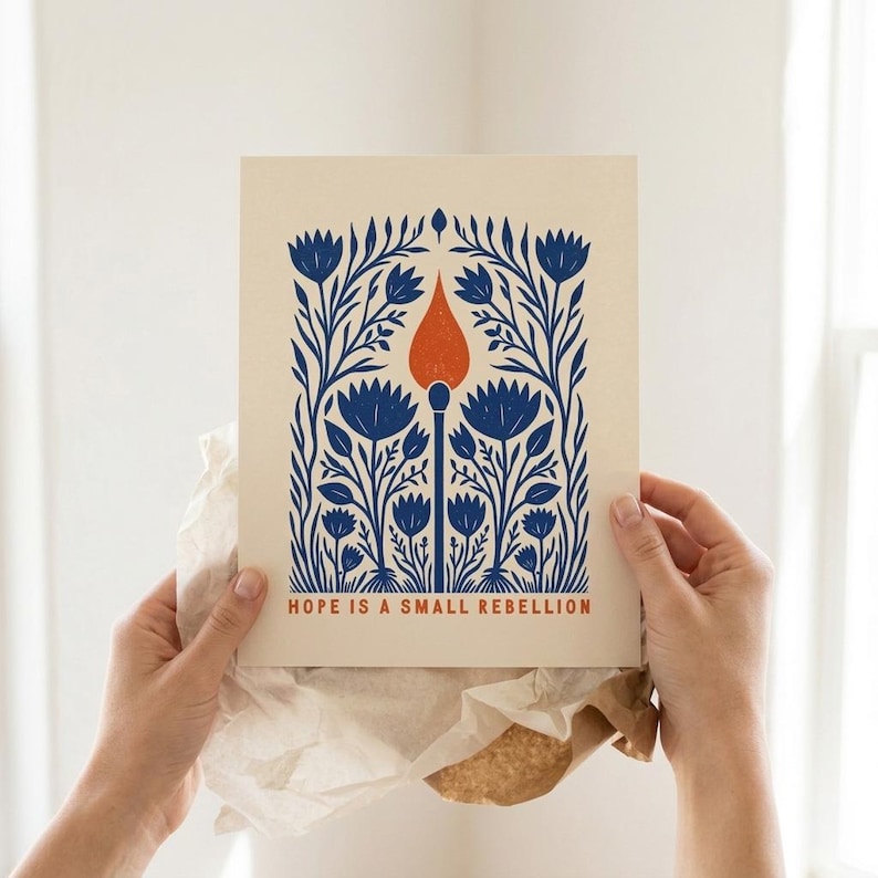 May include: A rectangular print on cream paper featuring a central orange matchstick surrounded by symmetrical blue floral patterns. The text "HOPE IS A SMALL REBELLION" is printed in orange at the bottom of the illustration in a clean, sans-serif font.