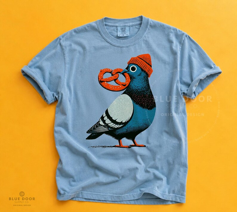 Pigeon shirt in ice blue Comfort Colors cotton on a mustard background, a New York pigeon in an orange beanie holding a pretzel