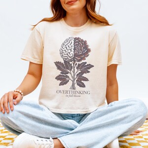 May include: Cream-colored t-shirt featuring a graphic of a brain and flower, with the text "OVERTHINKING in full bloom." The design is in shades of brown and gray. The person is wearing light blue jeans.