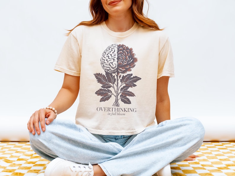 May include: Cream-colored t-shirt featuring a graphic of a brain and flower, with the text "OVERTHINKING in full bloom." The design is in shades of brown and gray. The person is wearing light blue jeans.