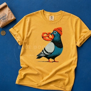 Pretzel shirt in mustard on cobalt with a soft pretzel bag prop, dopey NYC pigeon in an orange beanie