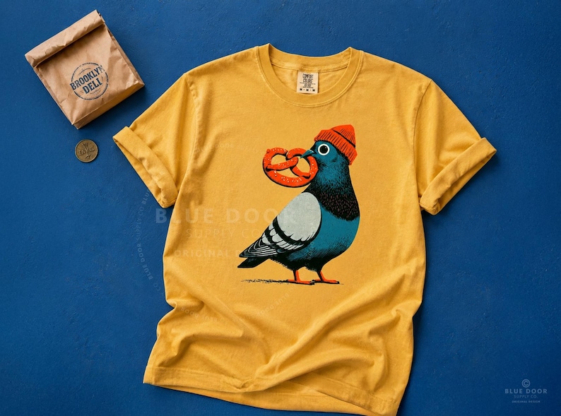 Pretzel shirt in mustard on cobalt with a soft pretzel bag prop, dopey NYC pigeon in an orange beanie